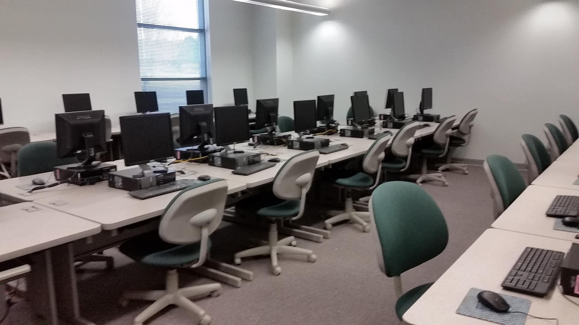 An empty computer lab with several workstations, each equipped with monitors, keyboards, and mice. The room has light-colored walls, green chairs, and a window with blinds on the left, allowing natural light to enter.