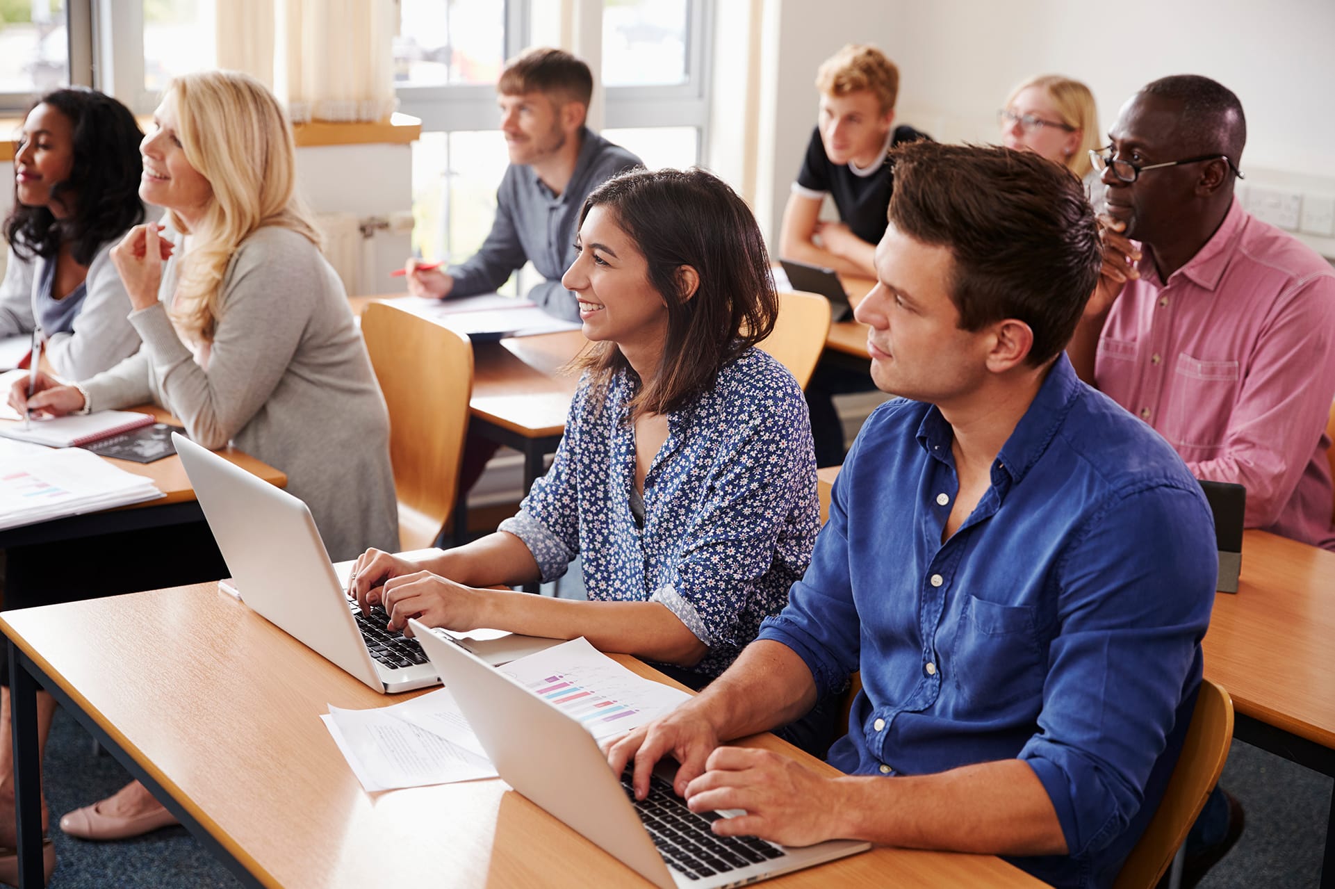 A diverse group of adults sits attentively in a classroom. Some are using laptops, while others have notebooks. They are facing forward, suggesting a lecture or discussion is taking place. The room is bright with natural light coming through the window.