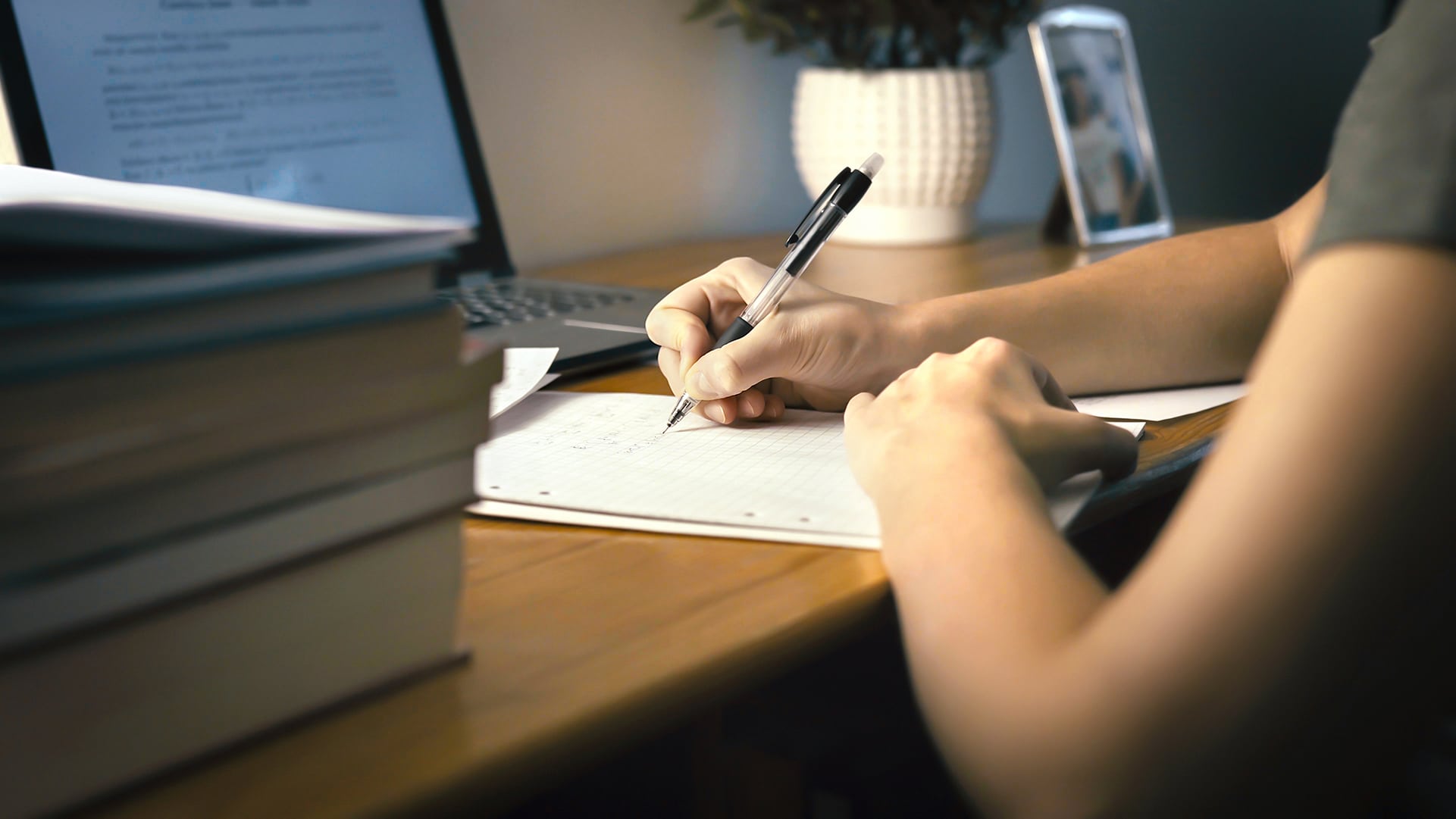 A person is sitting at a wooden desk, writing on lined paper with a pen. A laptop is open in the background, and several stacks of books are nearby. The lighting is warm, and a small potted plant is also visible on the desk.