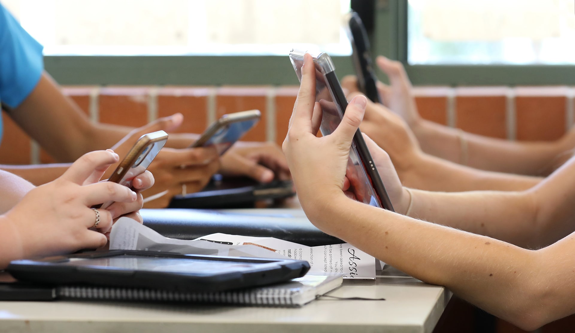 People sitting at a table, using smartphones. Some have notebooks and tablets on the table. A brick wall and window are in the background, indicating an indoor setting. The focus is on their hands and devices.