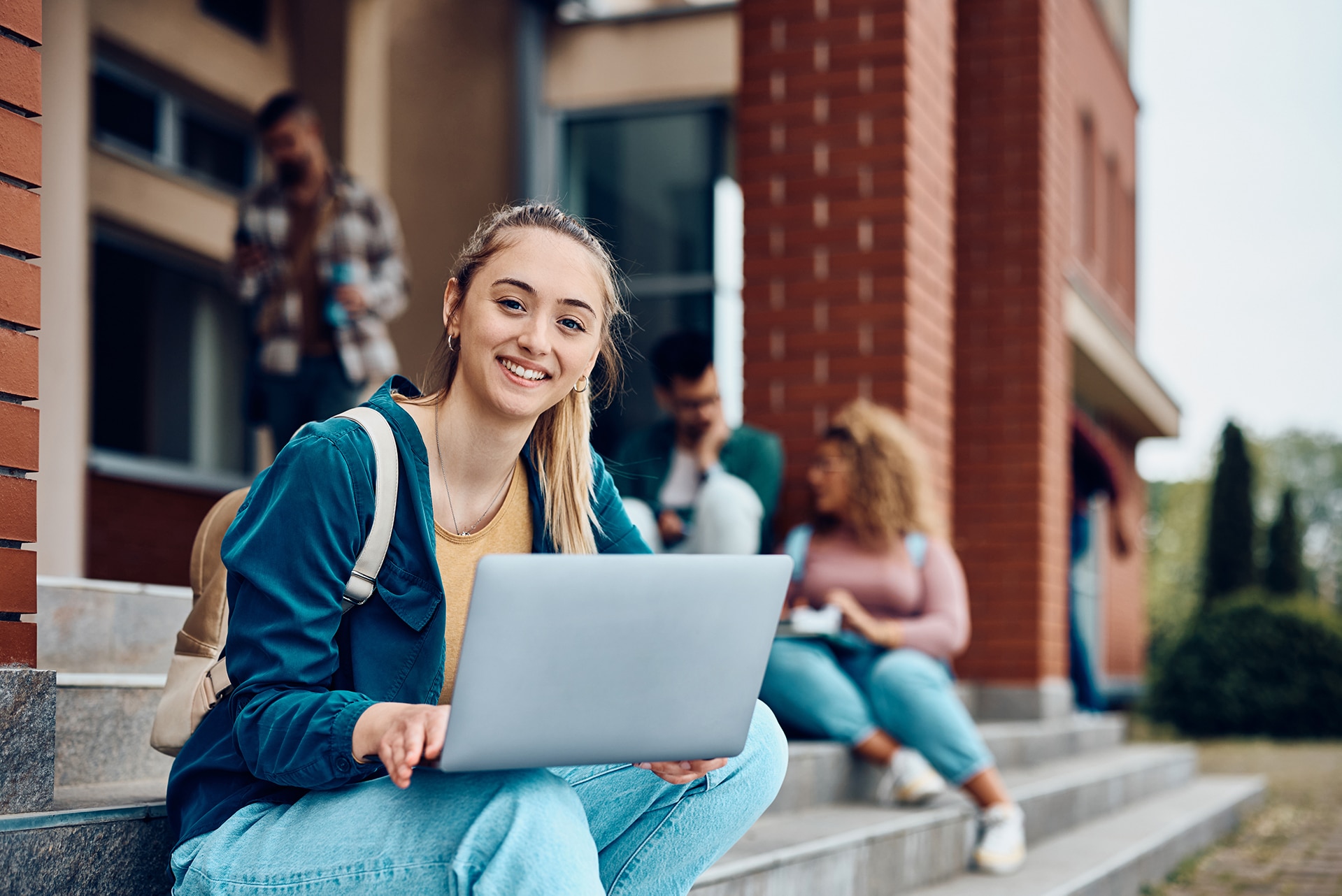 A young woman sitting on outdoor steps, smiling and using a laptop. She wears a backpack and blue jacket. In the background, three people sit on steps, engaged in conversation. The setting appears to be a school or university.