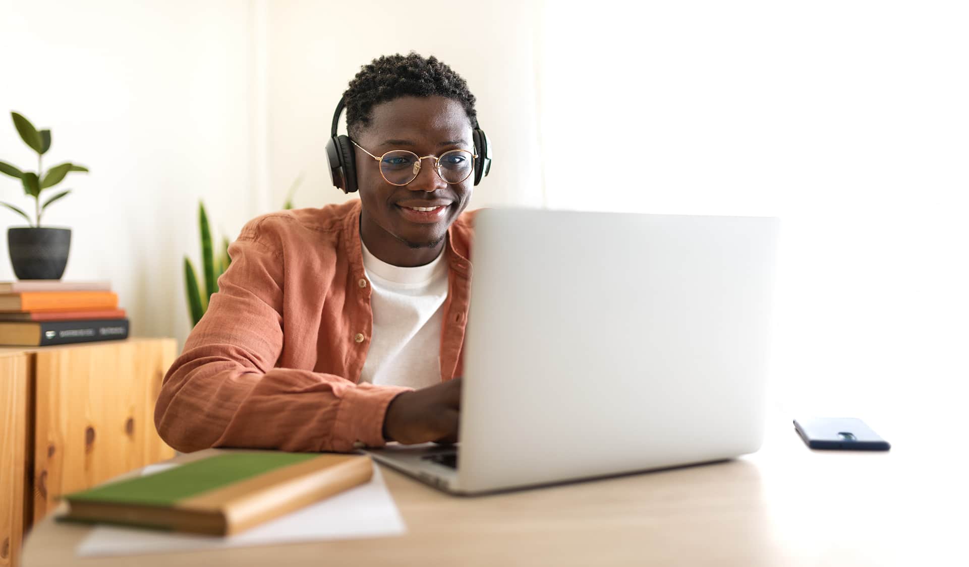 A person wearing headphones and glasses is smiling while using a laptop at a desk. Books and a plant are on the desk, and a phone is nearby. The setting is bright and inviting.