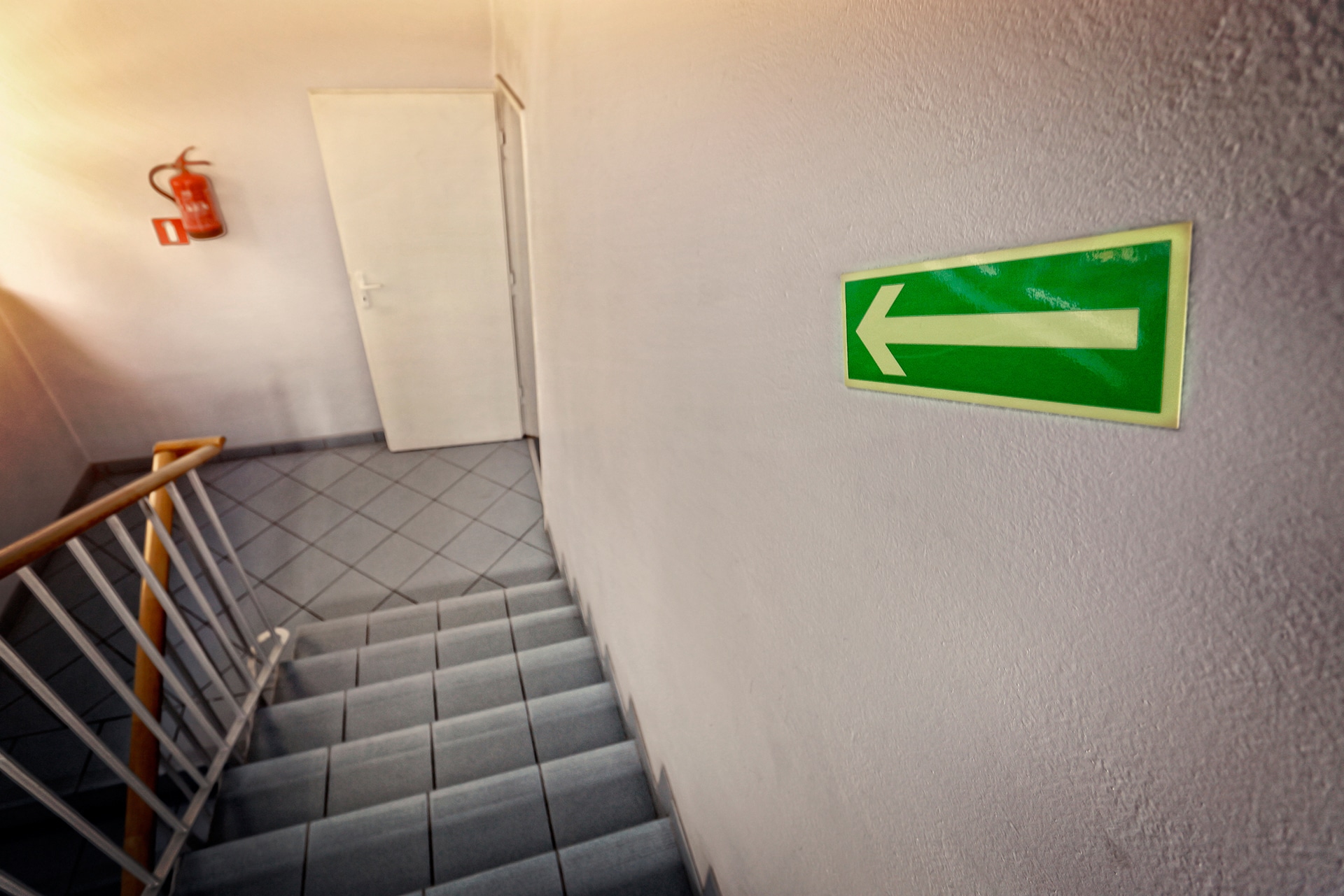 A stairway with light gray tiles and a white door at the top. A green emergency exit sign with a left-pointing arrow is on the wall near the stairs. A wooden handrail lines the stairs, and a fire extinguisher is mounted on the wall.