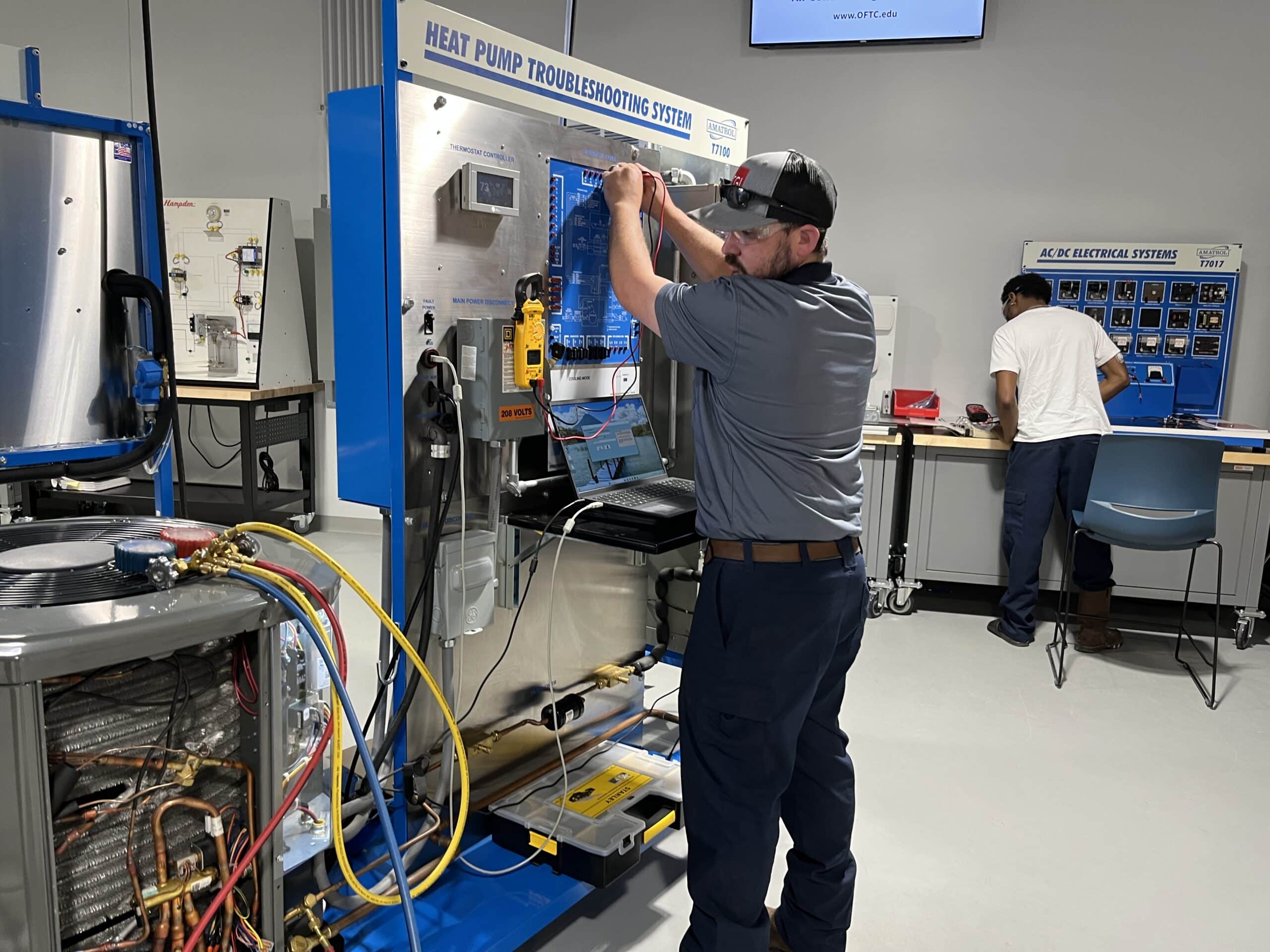 Two men are working in a lab equipped with HVAC training devices. One is adjusting controls on a heat pump troubleshooting station, while the other is seated at a workstation in the background. Hoses and gauges can be seen on the left side of the image.