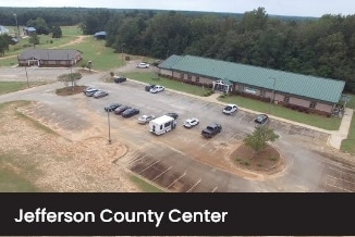 Aerial view of Jefferson County Center featuring a large green-roofed building, several parked cars, and a surrounding forested area. A smaller building is visible in the background.