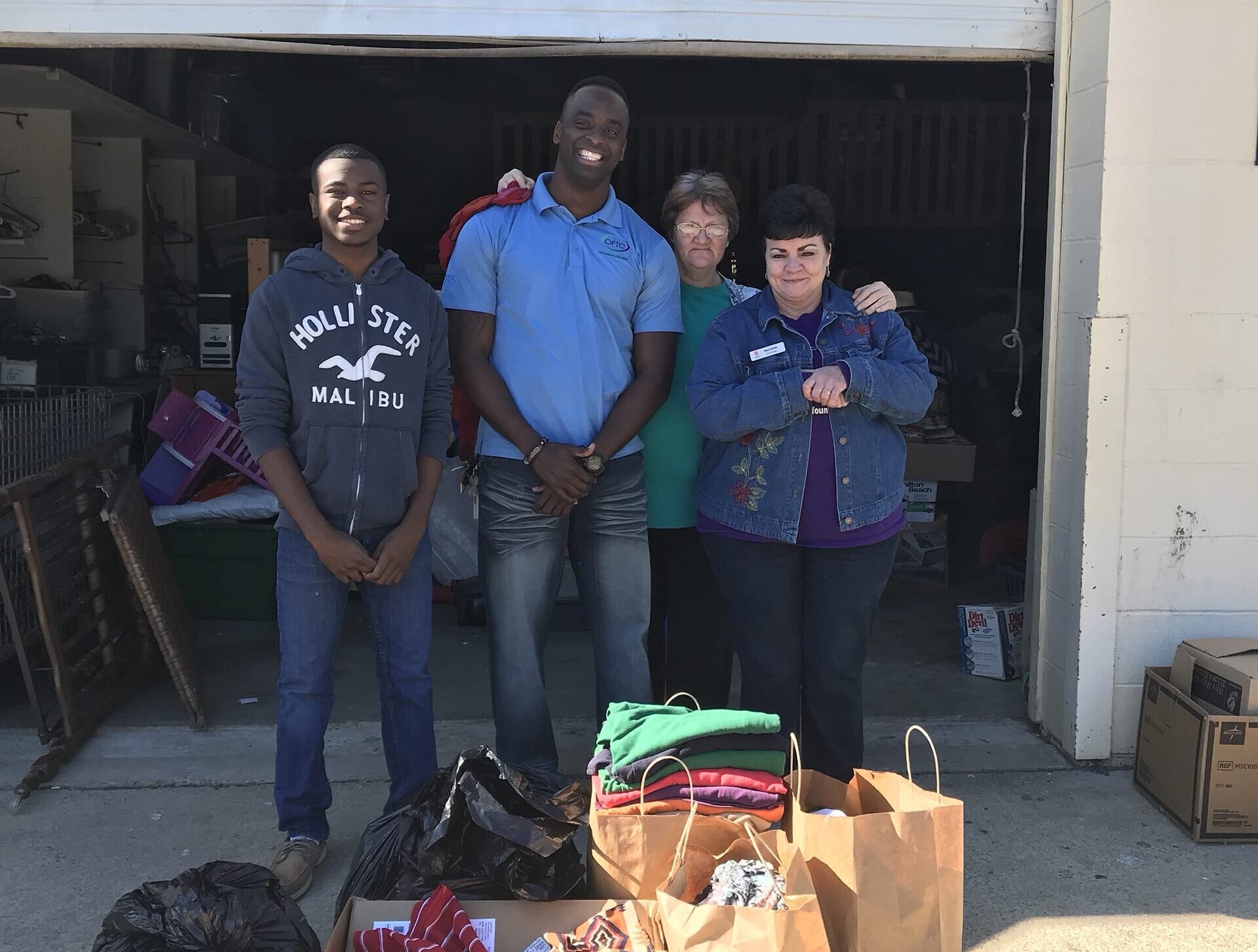 A group of four people stands in front of a garage. They are smiling and have their arms around each other. In front of them, there are bags and a box filled with clothing and other items.