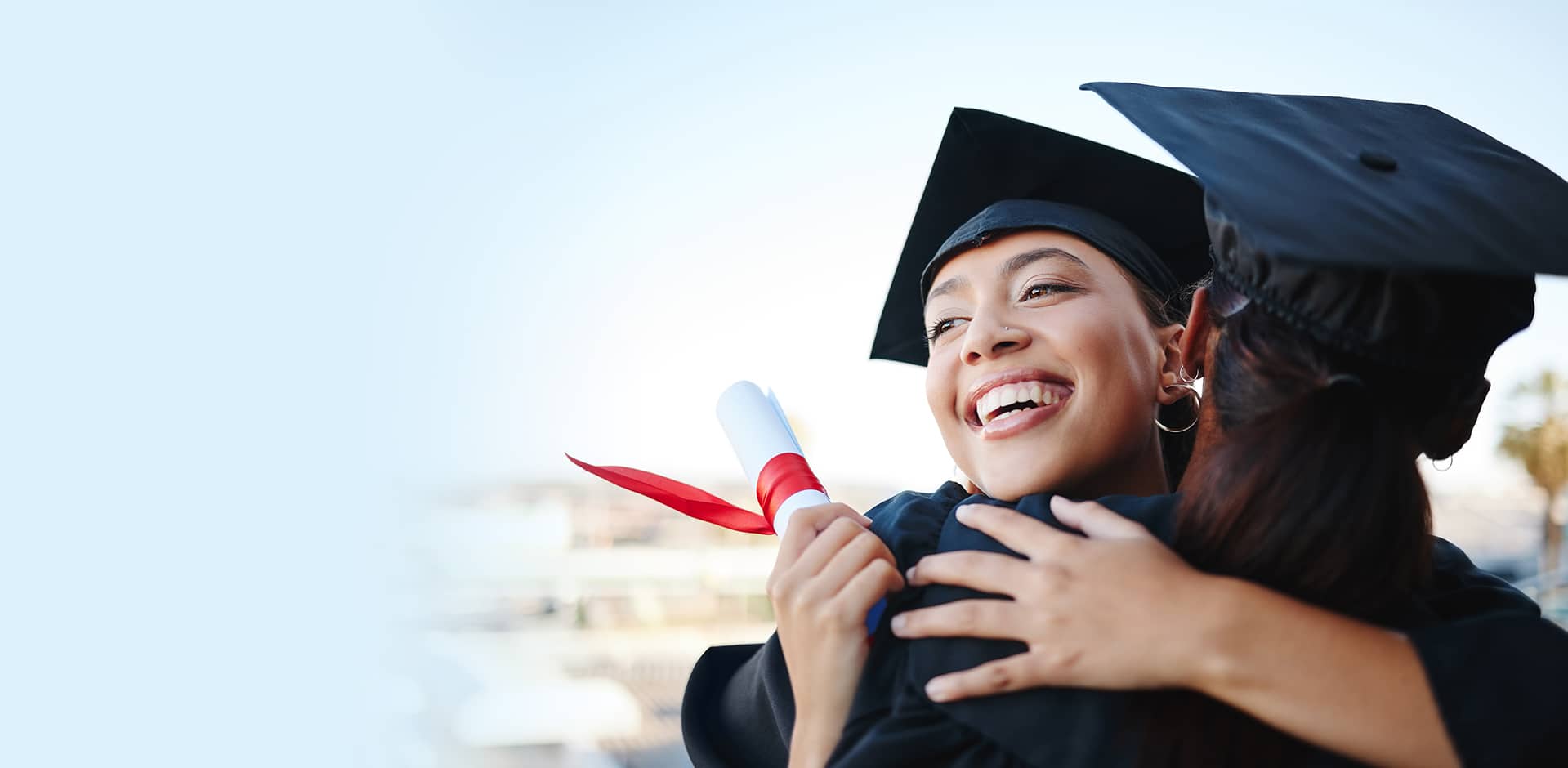 Two graduates in caps and gowns embrace joyfully. One holds a diploma tied with a red ribbon. The background is softly blurred, emphasizing their happiness and achievement.