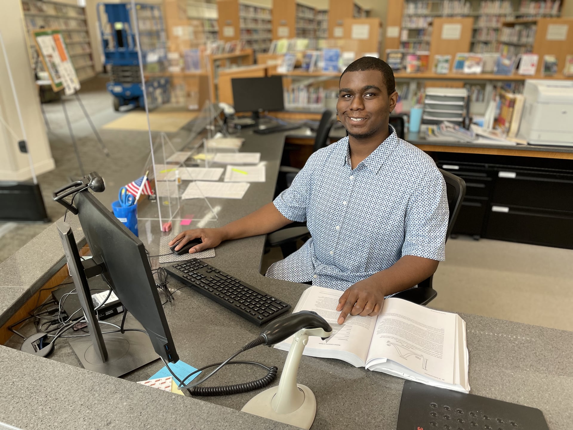 A person sits at a library desk with an open book and a computer. They are smiling, wearing a patterned short-sleeve shirt. Bookshelves filled with books are visible in the background. Office supplies and a flag are on the desk.