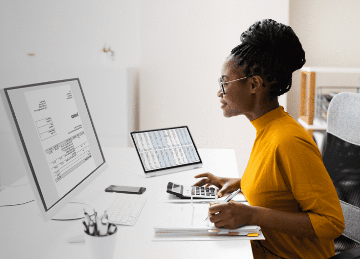 A woman in a yellow top sits at a desk, working on a computer with a document displayed on the monitor. She holds a calculator, with a tablet and smartphone nearby. The setting is a bright office space with modern decor.