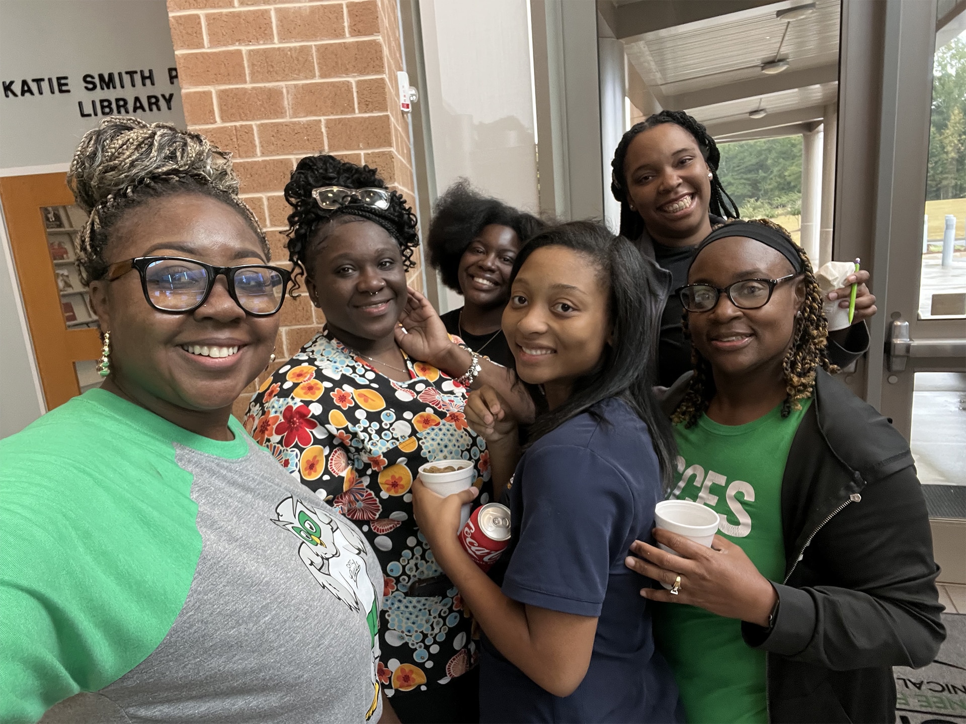 A group of six smiling women pose together indoors. They are holding drinks and standing near a doorway labeled KATIE SMITH P LIBRARY. The women are wearing casual clothing including a colorful floral dress and a green graphic t-shirt.