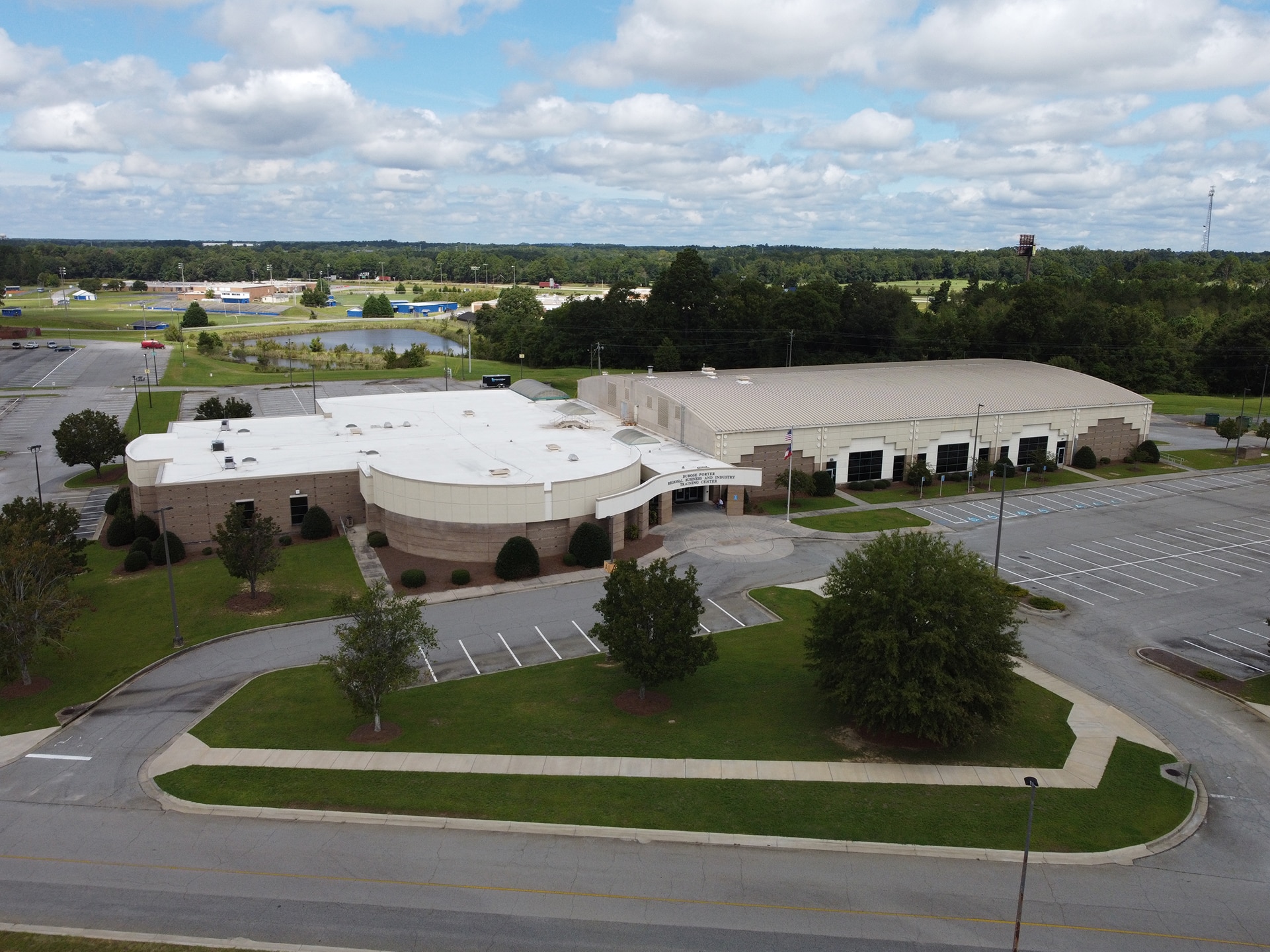 Aerial view of a large, modern building with a white roof and rounded sections, surrounded by parking lots and green spaces. There are several trees and a few cars in the parking area. The sky is partly cloudy, with green fields in the background.
