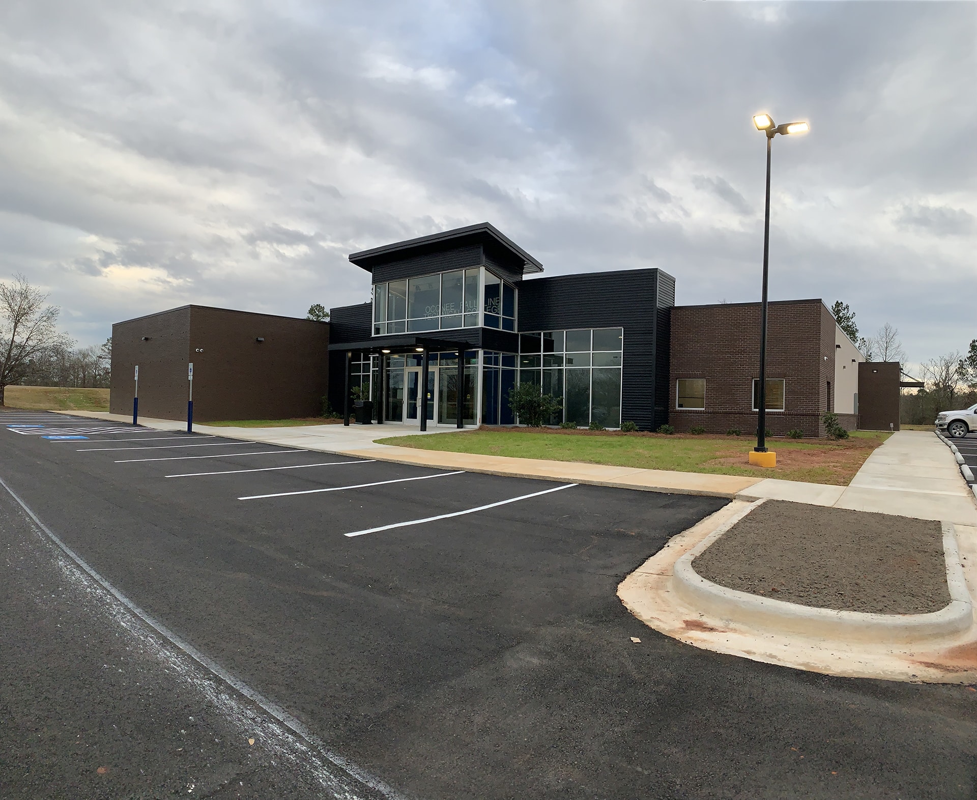 A modern, one-story building with large glass windows and a dark facade sits empty. The surrounding area features a paved parking lot with a few streetlights and some parked cars. The sky is overcast.