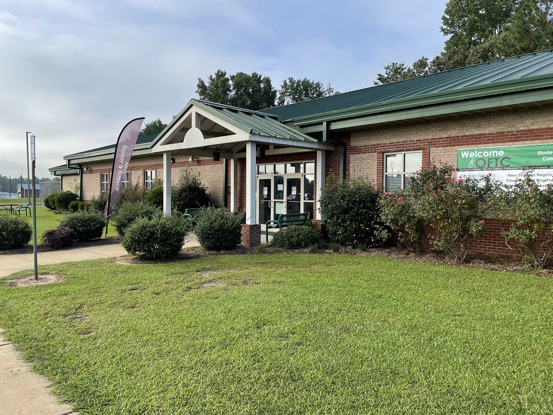 A brick building with a green metal roof, featuring a main entrance under a small portico. There's a sign reading Welcome and lush green grass and shrubbery surrounding the building. A few trees are visible in the background.