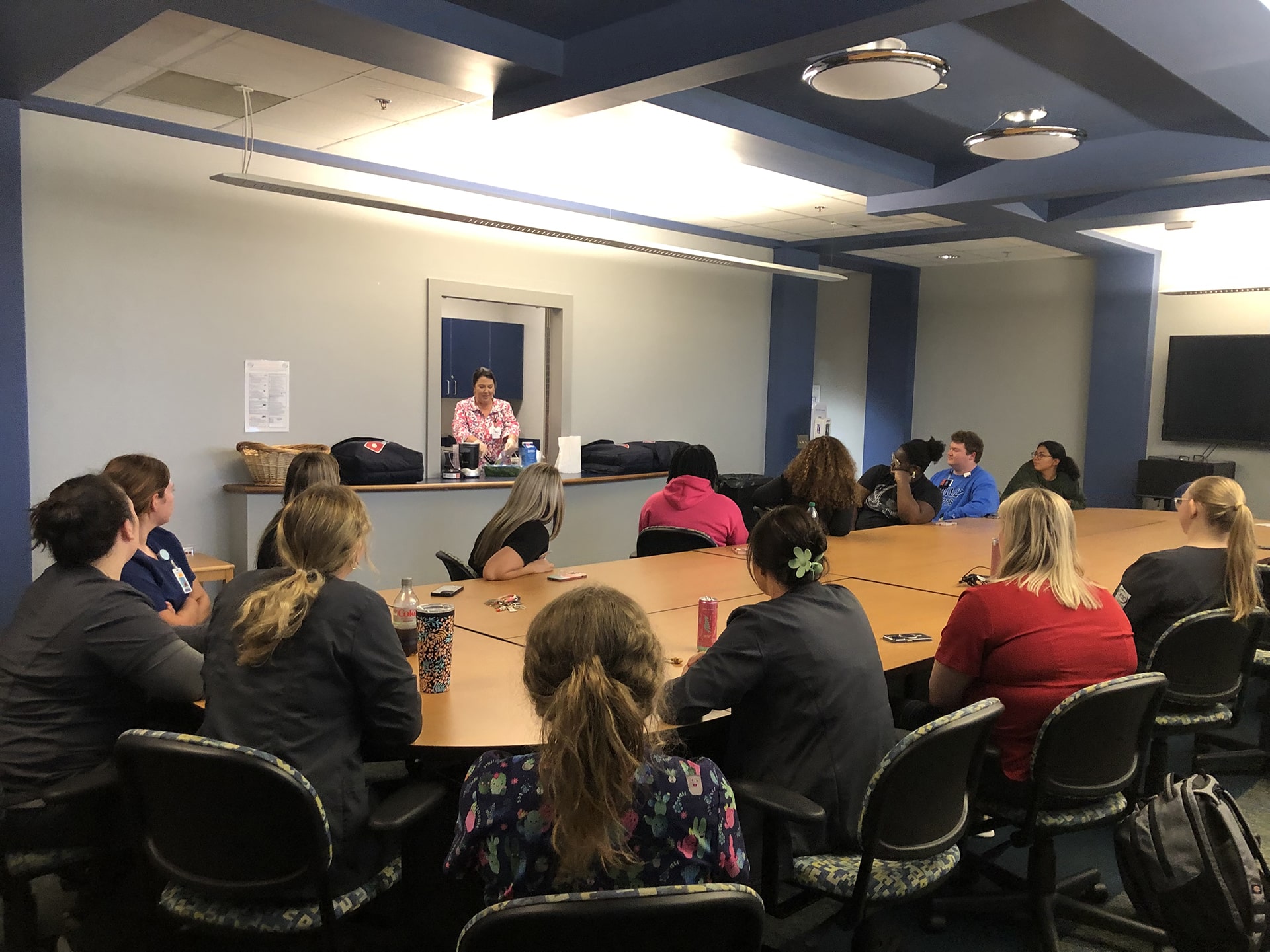 A group of people sit around a large conference table listening to a person speaking at the front of the room. The room has blue walls, a ceiling with recessed lights, and a flat-screen TV on the wall. The speaker stands near a podium.
