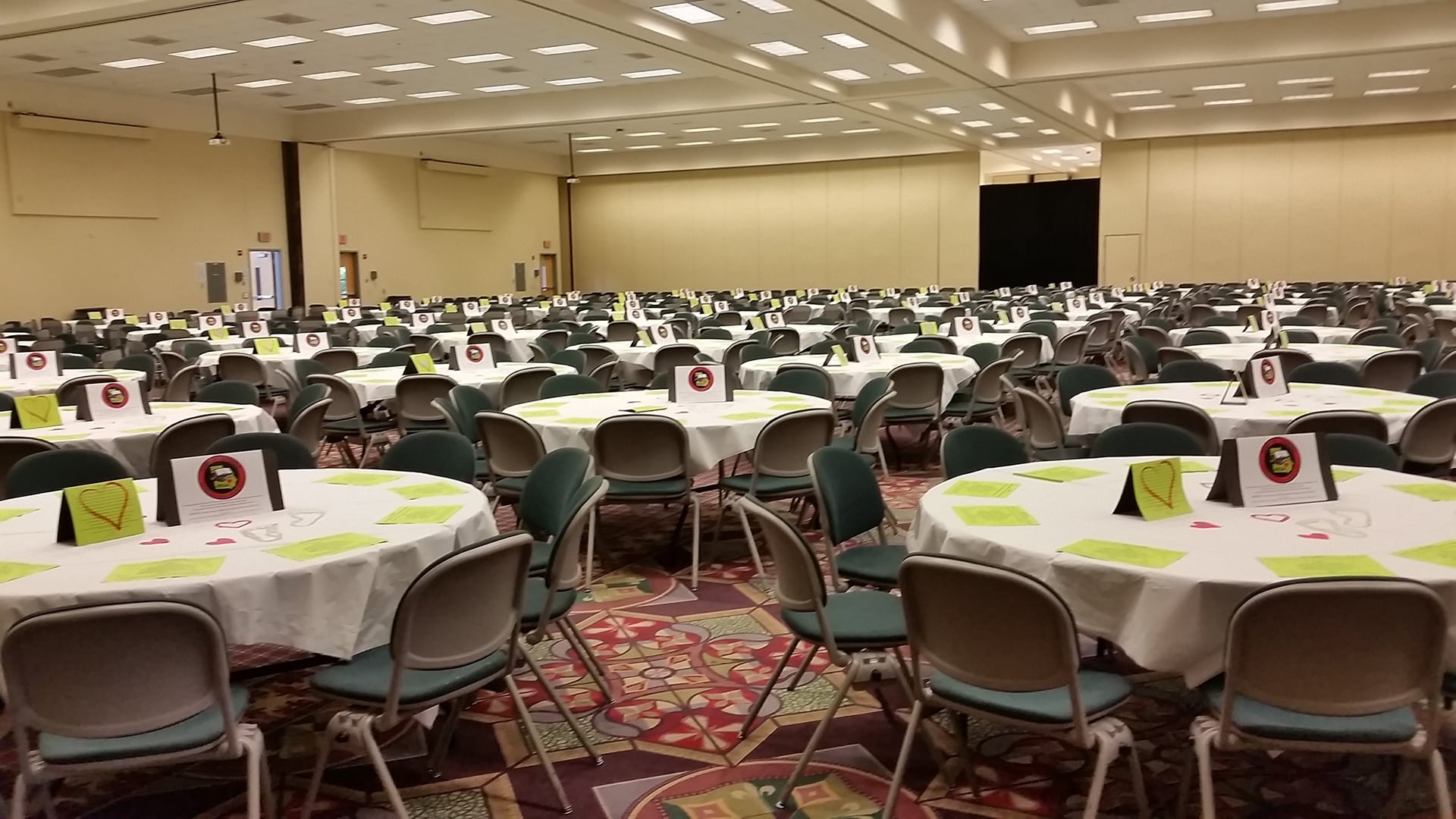 A large banquet hall with numerous round tables covered with white tablecloths. Each table is set with chairs and decorated with centerpieces and paper items on a patterned carpet under bright overhead lights. The room appears empty.
