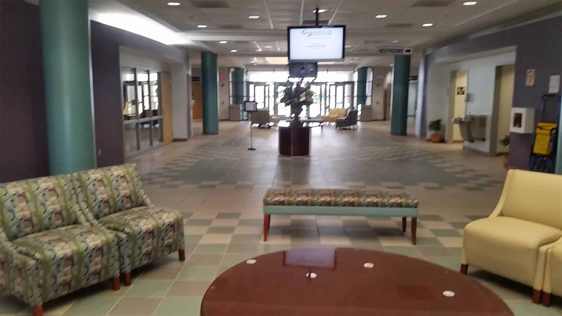 Hospital waiting area with patterned chairs, a bench, and a round table. A TV screen hangs from the ceiling, and a reception area with signs to various departments is visible. The floor is tiled in green and beige.