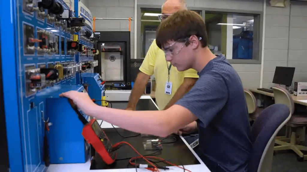 A young person wearing safety goggles operates electronic equipment at a workstation, adjusting settings on a panel with various switches and dials. An older person stands behind, observing the process. They are in a lab or workshop setting.