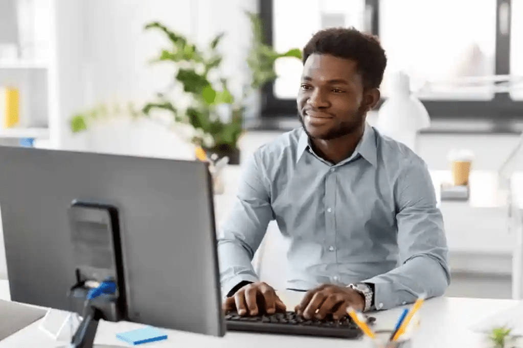 A man in a light gray shirt is sitting at a desk, typing on a keyboard. He is smiling while looking at a large monitor. The office setting includes plants and bright natural light from the windows.