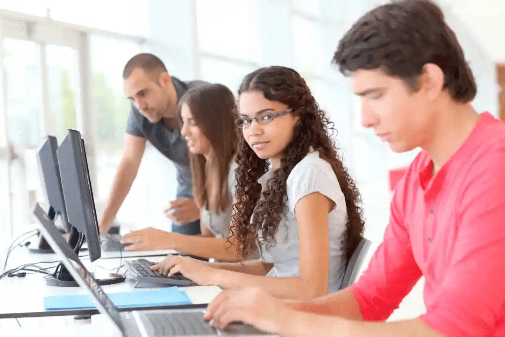 Four young adults work on computers in a bright room. Two women and two men are focused on their screens, collaborating in pairs. They are seated at a shared table with multiple monitors, suggesting a tech or study environment.
