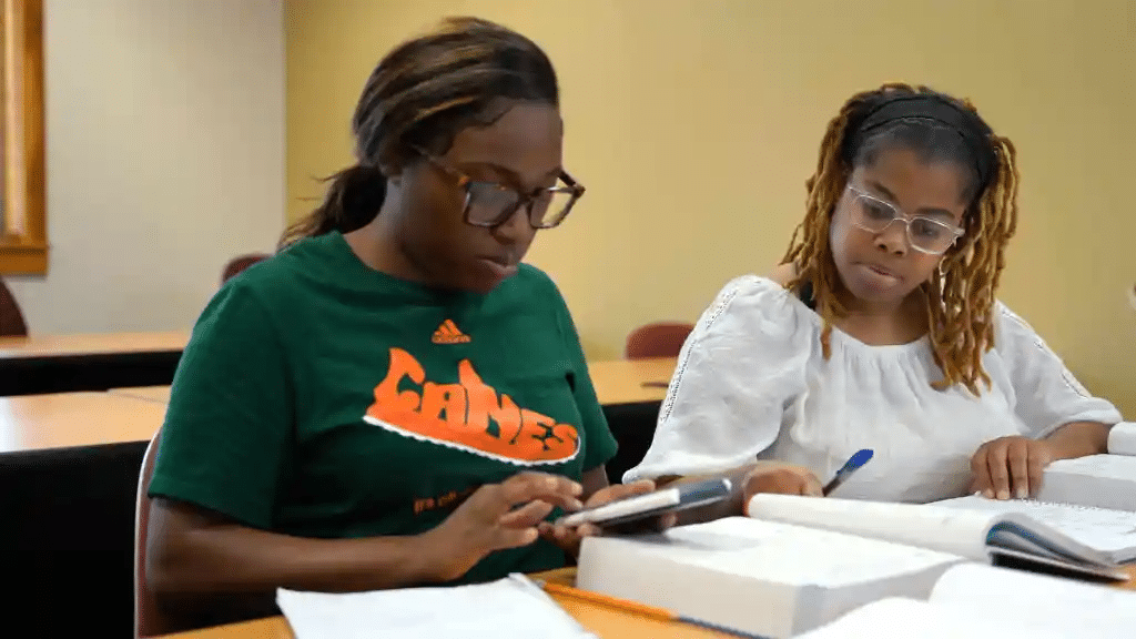 Two people sitting at a classroom table, studying. One wears a green shirt and glasses, using a calculator. The other, in a white blouse and glasses, is holding a pen, looking at a book. Both are focused on their work.