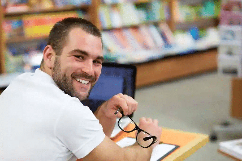 A man with a beard, wearing a white shirt, smiles while holding glasses in one hand. He is seated at a desk with a laptop in a library or bookstore, with shelves of books blurred in the background.