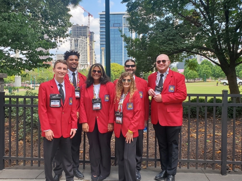 Six people standing outside on a paved area in front of a black fence, wearing matching red blazers with badges. They are smiling, with trees and tall buildings in the background, under a partly cloudy sky.