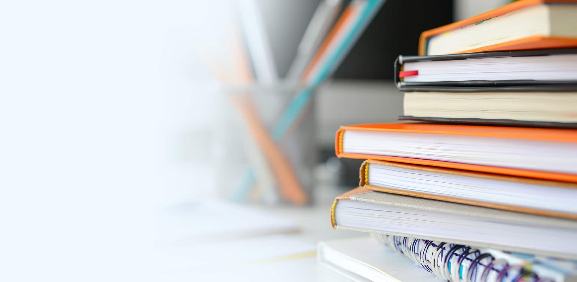 A stack of colorful books and notebooks is placed on a desk. The background is blurred with faint outlines of more books and stationery. The image conveys a study or learning environment.