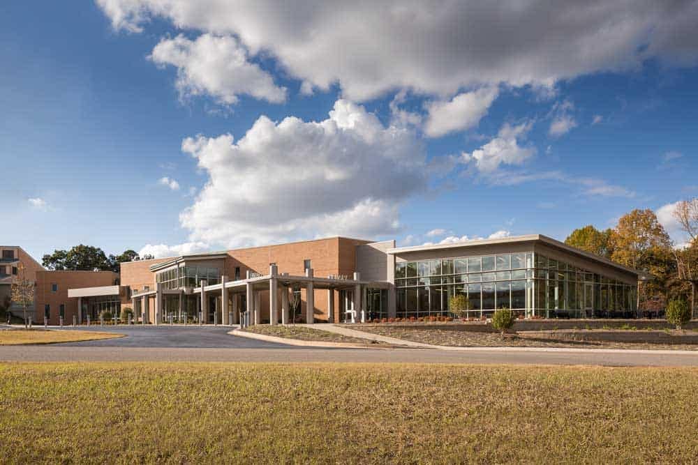 Modern building with a mix of brick and glass architecture, featuring a covered entrance and large windows. The sky is partly cloudy, and the surrounding area includes grass and trees.