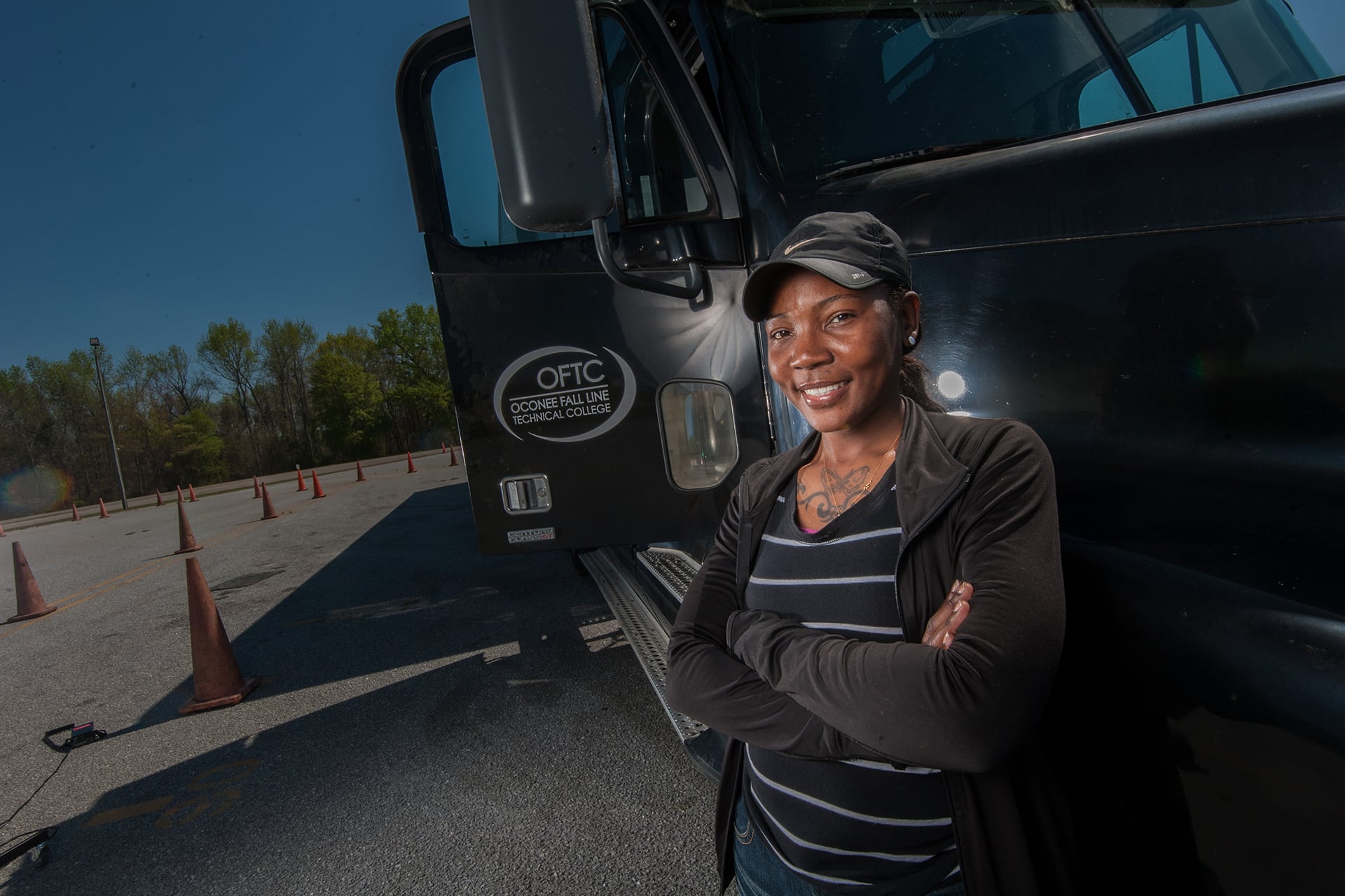 The alt text has no quotation marks: A person stands confidently with arms crossed in front of a large black truck. They are wearing a black cap and a striped shirt. Orange traffic cones line the road beside the truck. Trees and a clear blue sky are visible in the background.