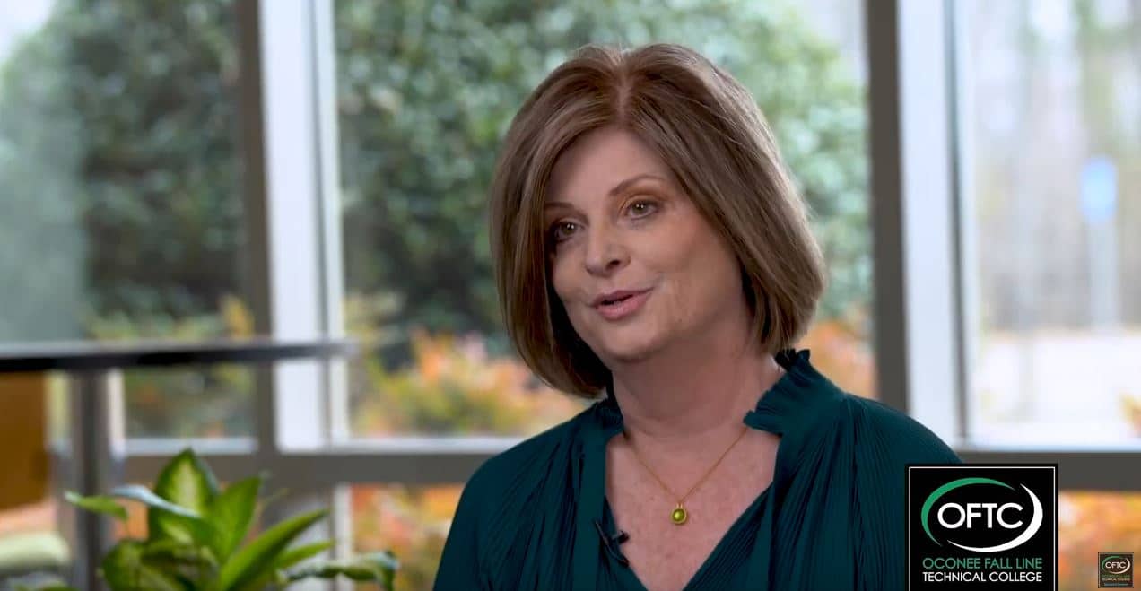 A woman with short brown hair and a teal blouse is speaking in an indoor setting. There's a backdrop of large windows showing trees and plants. A logo in the bottom right corner reads OFTC Oconee Fall Line Technical College.