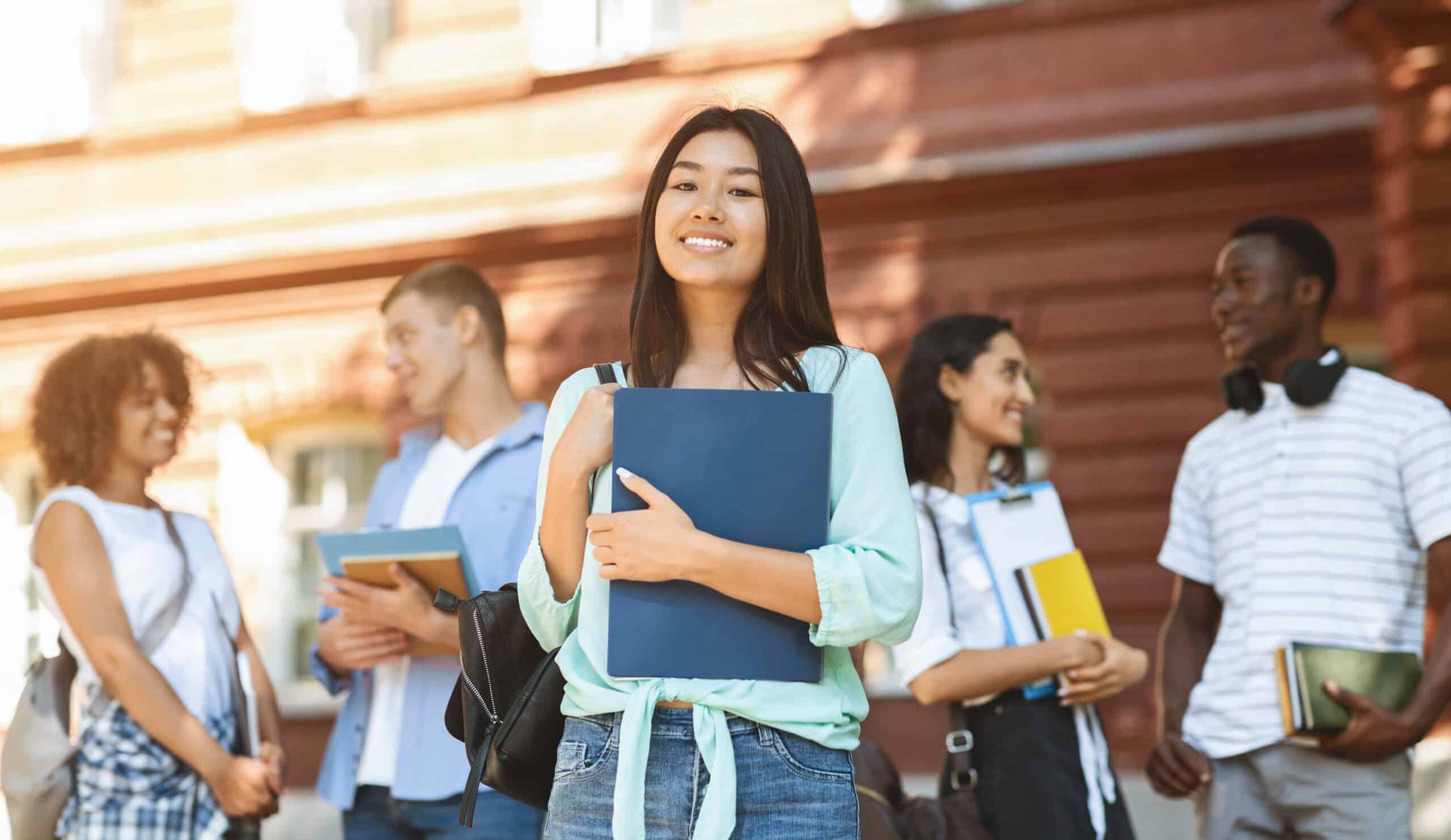 A young woman holding a blue folder stands smiling in front of a large brick building. In the background, four other students are mingling, holding books and backpacks, and appearing engaged in conversation.
