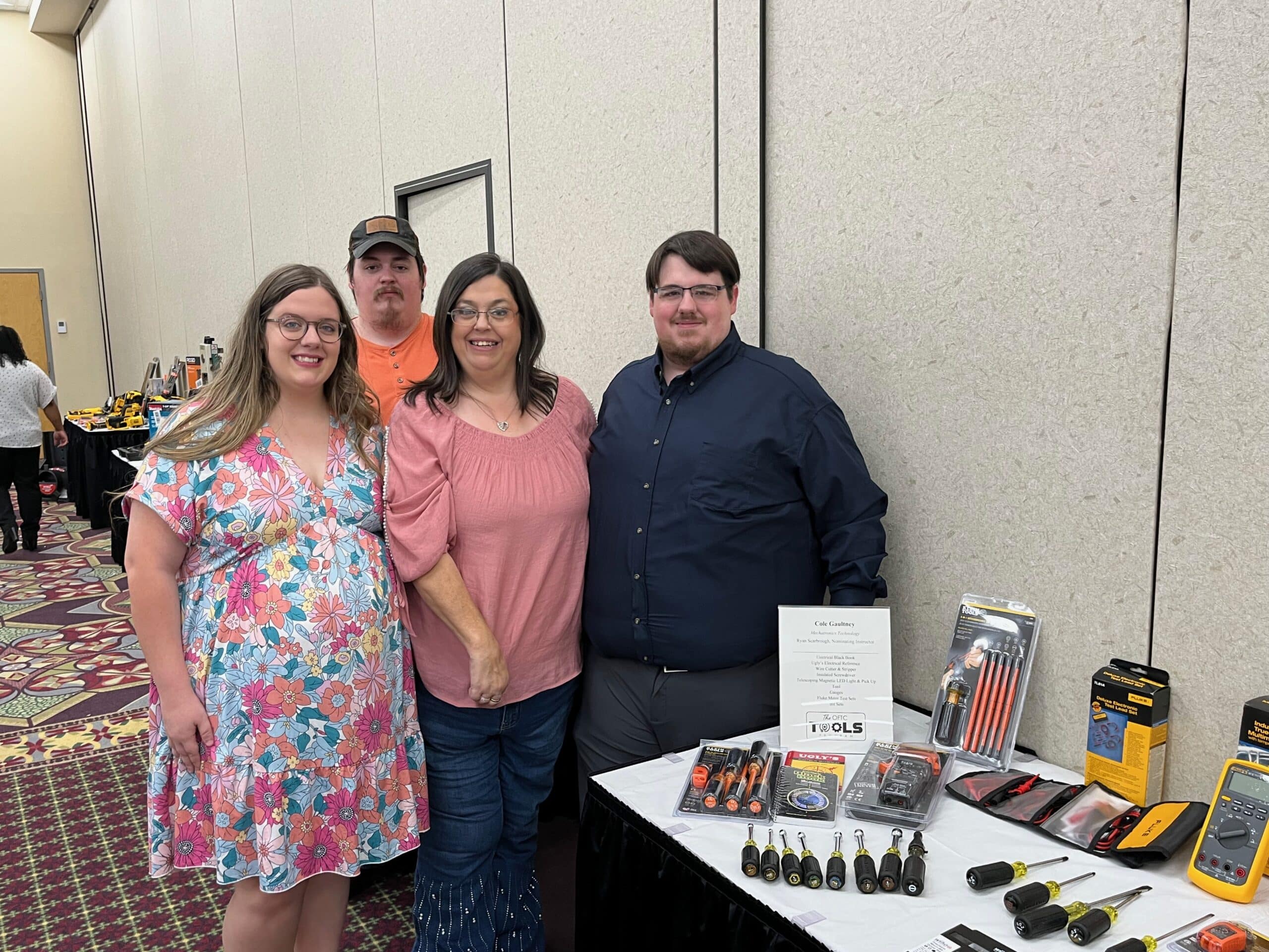 Four people stand together beside a display table with various tools and equipment in a conference room. The table has screwdrivers, multimeters, and other packaged tools. The group smiles at the camera.