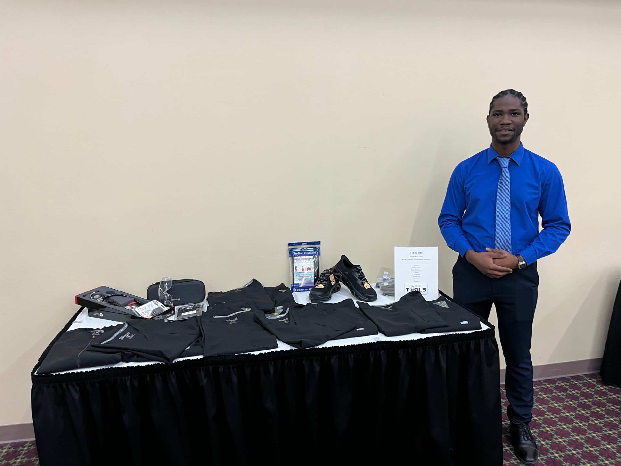 A man in a blue shirt and tie stands beside a display table showcasing black athletic clothing, shoes, and accessories, all neatly arranged against a beige wall with a patterned carpet.