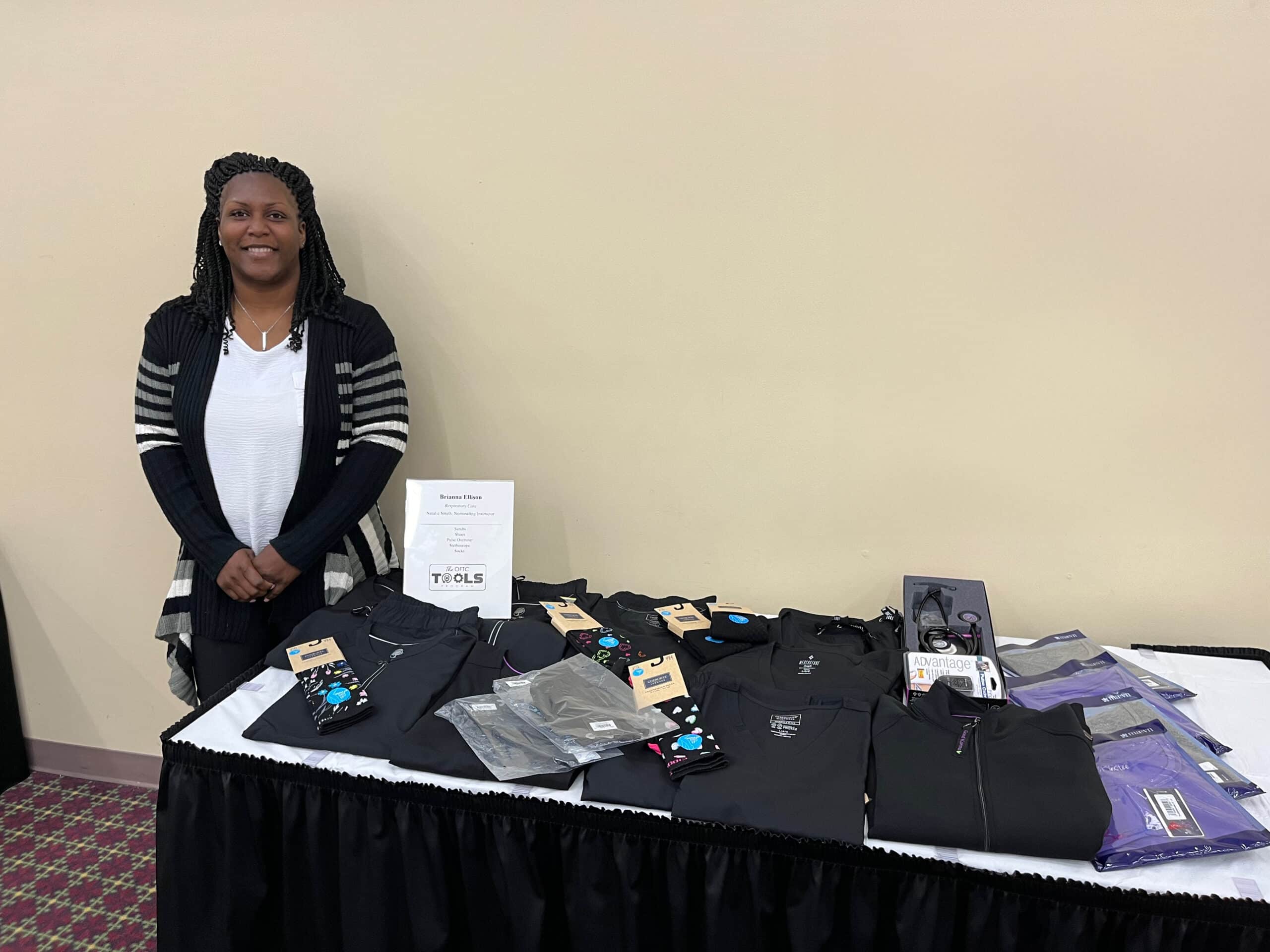 A woman stands beside a display table featuring neatly arranged black clothing, socks, and accessories. The table has a white sign and is set against a beige wall in an indoor setting.