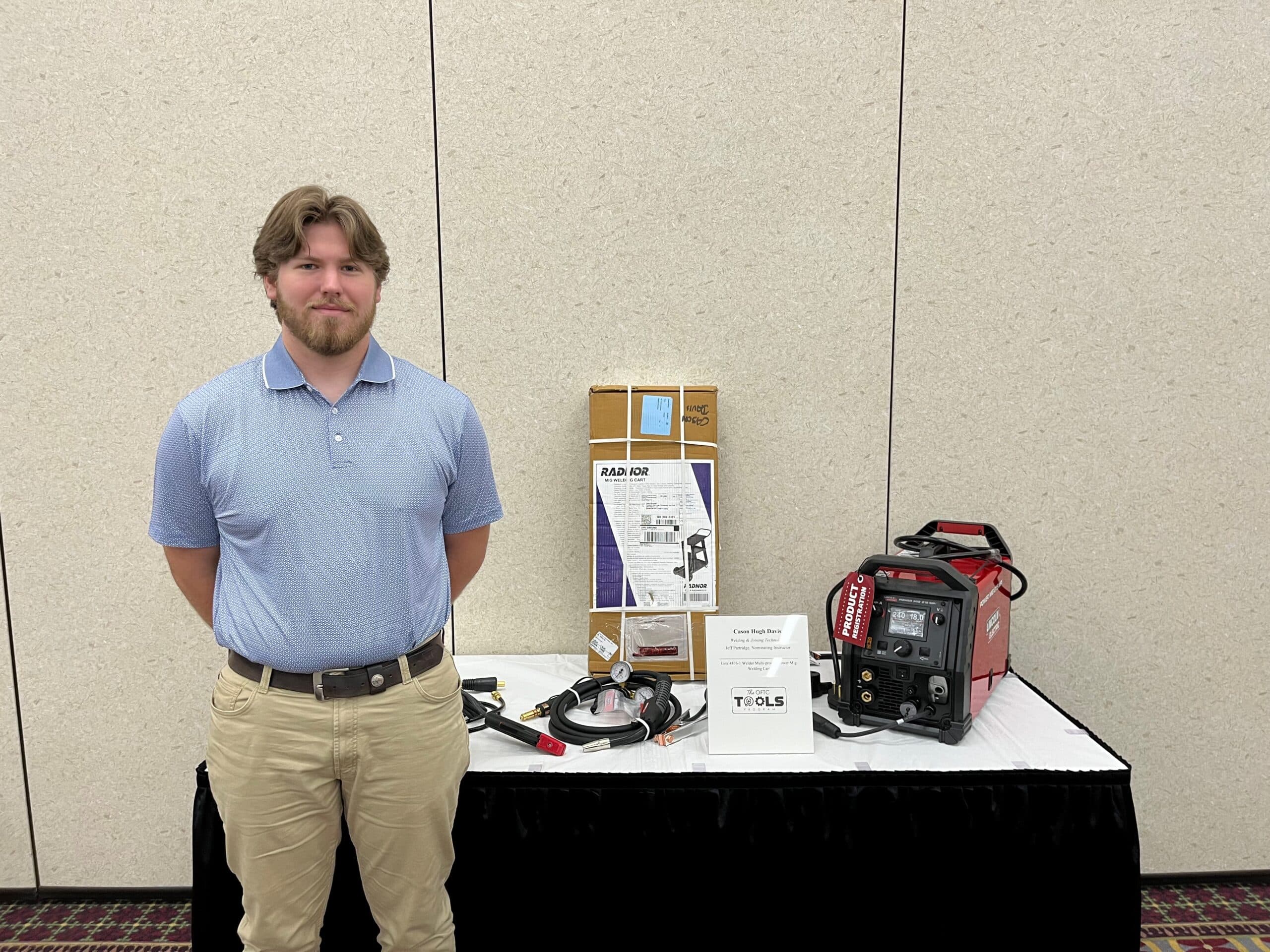 A man with light brown hair and a beard stands beside a table displaying welding equipment, tools, a boxed item, and a small sign, all set against a beige wall.
