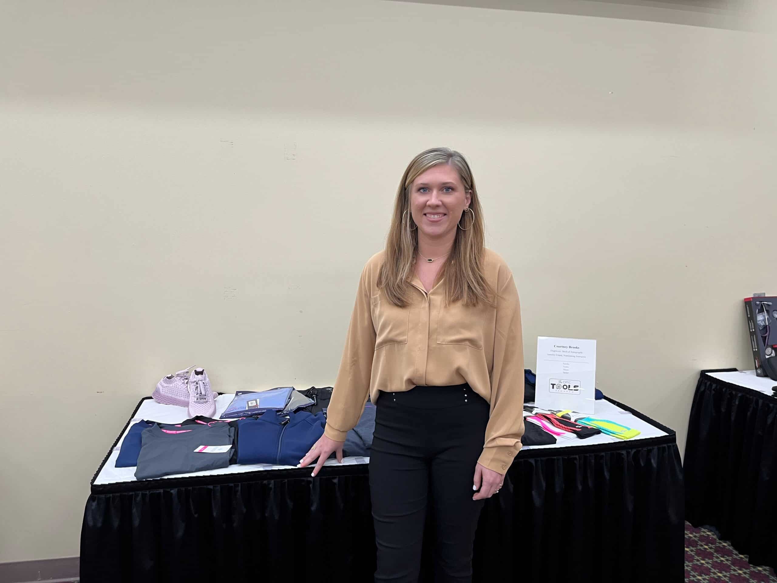 A woman with long blonde hair, wearing a tan blouse and black pants, stands in front of a display table with clothing and accessories on it. The background shows cream-colored walls and more display tables.