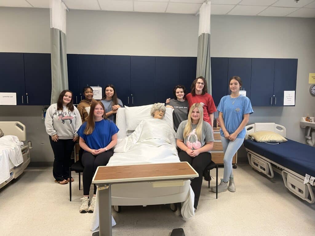 Nine students pose around a hospital bed with a medical training mannequin in a classroom setting, surrounded by beds and blue cabinets.