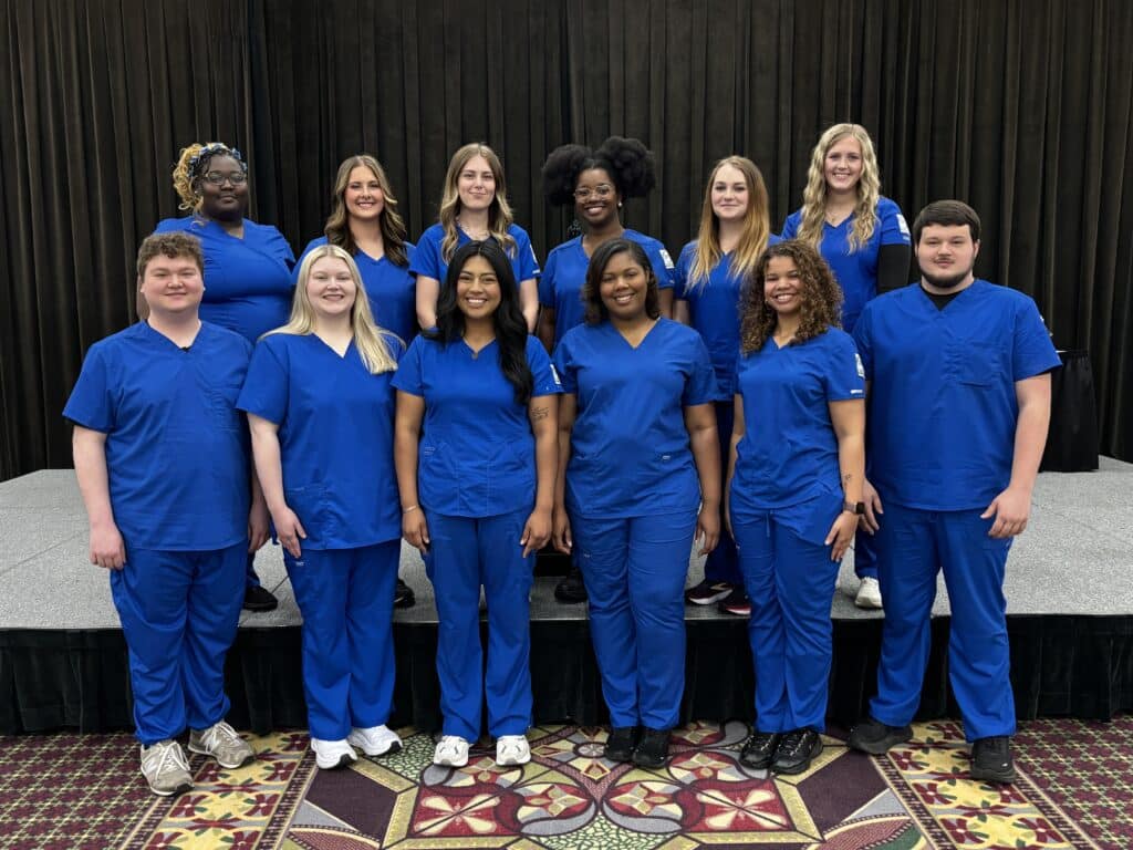 Twelve people in blue medical scrubs stand together on a carpeted floor and raised platform, posing and smiling for a group photo in front of a dark curtain.