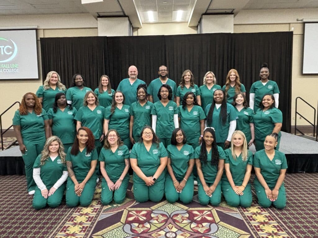 A group of 24 people wearing matching green scrubs pose together in rows on a patterned carpet in front of a black stage and a Falling Line Technical College sign. The setting appears to be indoors, possibly an event or graduation.
