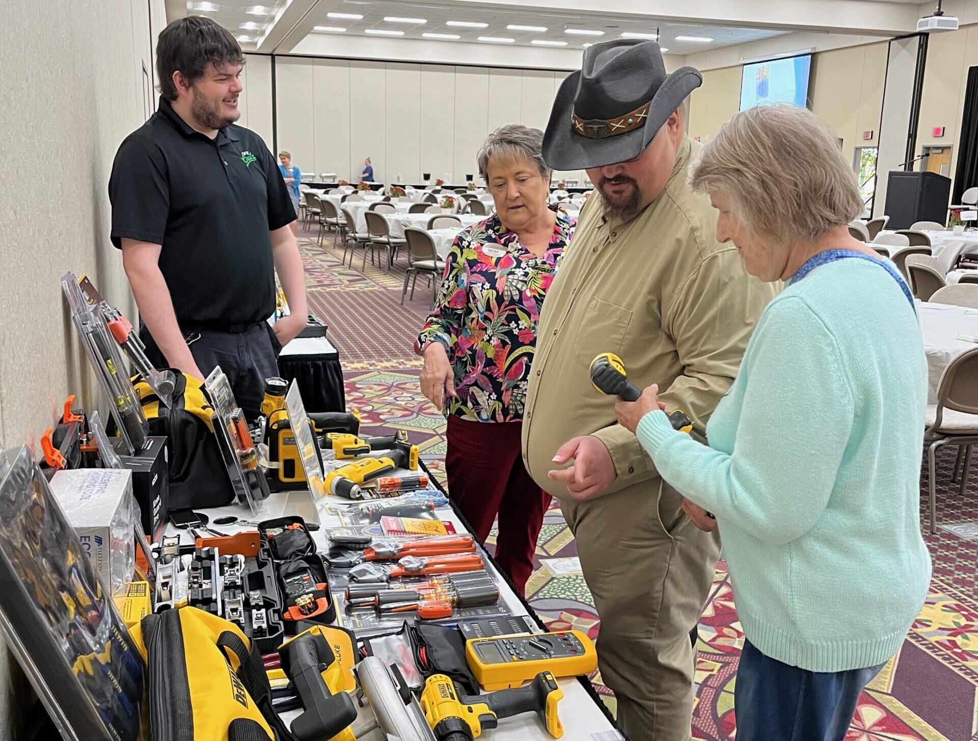 Three adults examine a display of power tools and equipment on a table while a vendor stands by and explains the products in a large well-lit conference room with many empty tables and chairs.