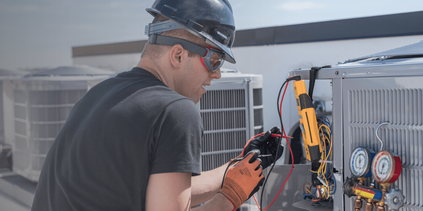 A technician wearing a hard hat, safety glasses, and gloves uses electrical testing equipment to check an air conditioning unit on a rooftop.