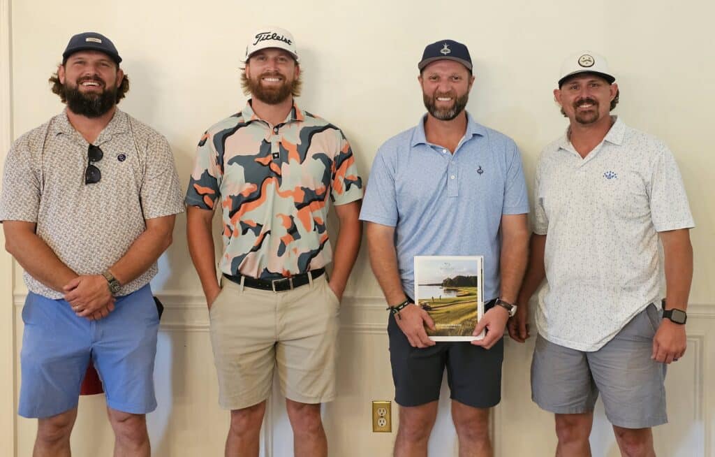 Four men stand side by side indoors, smiling at the camera. They wear casual golf attire, including polo shirts, shorts, and caps. One man holds a plaque or award. The background is a plain light-colored wall.