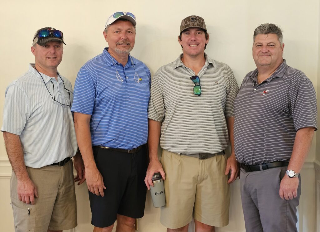 Four men stand side by side against a light-colored wall, all wearing polo shirts, shorts or pants, and casual hats. One man holds a gray Tumbler cup and they are smiling at the camera.
