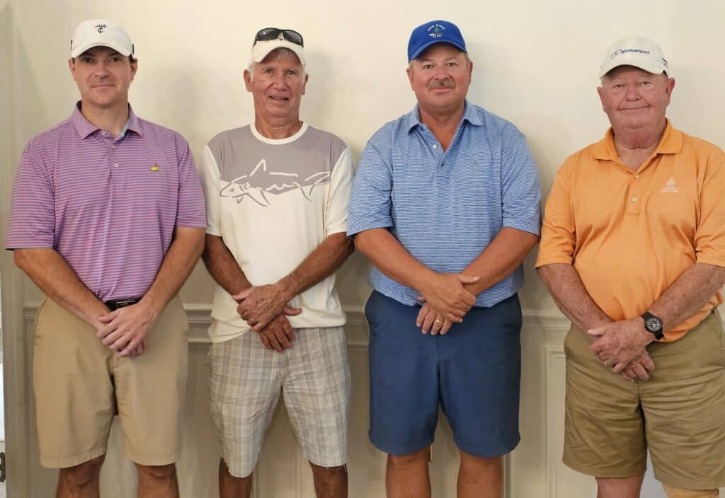 Four older men stand side by side indoors, each wearing a golf cap and casual golf attire—striped or patterned polo shirts and shorts—posing and smiling slightly in front of a plain, light-colored wall.