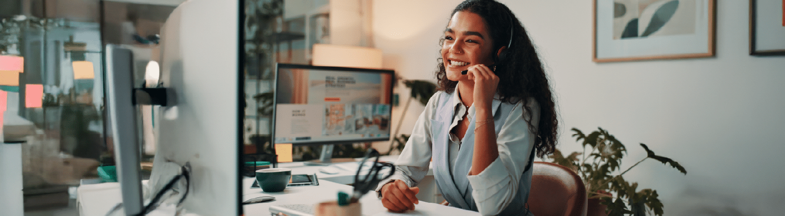 A woman wearing a headset sits at a desk and smiles while talking on the phone in a modern office with a computer, plants, and wall art visible in the background.