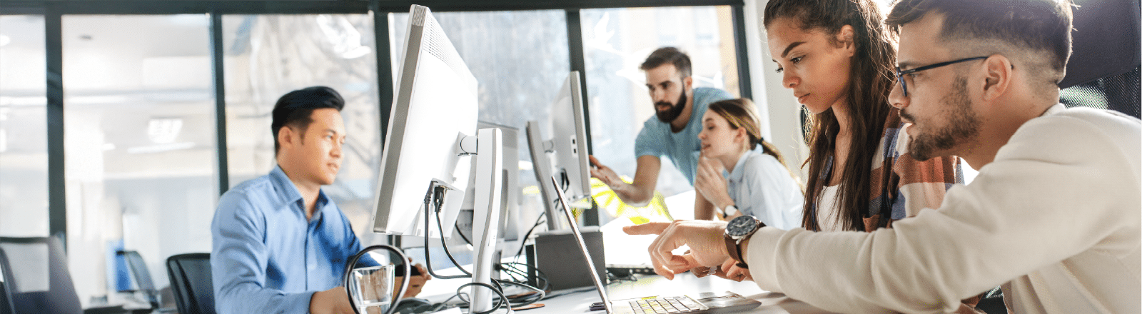Five young adults collaborate in a modern office, working at computers and discussing a project. In the foreground, two people focus on a monitor while others engage in conversation in the background.
