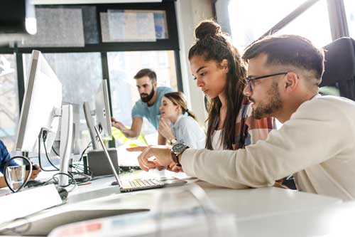 Two young adults work together at a computer in a modern office with other colleagues collaborating and talking in the background. Sunlight streams through large windows creating a bright productive atmosphere.