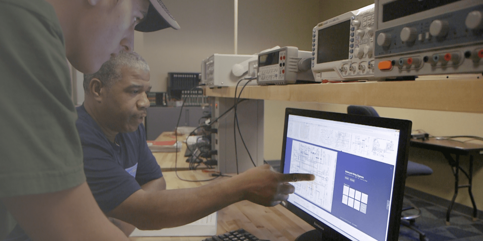 Two people in a lab sit at a desk looking at technical diagrams on a computer monitor. One person points at the screen while electronic equipment is visible on shelves behind them.