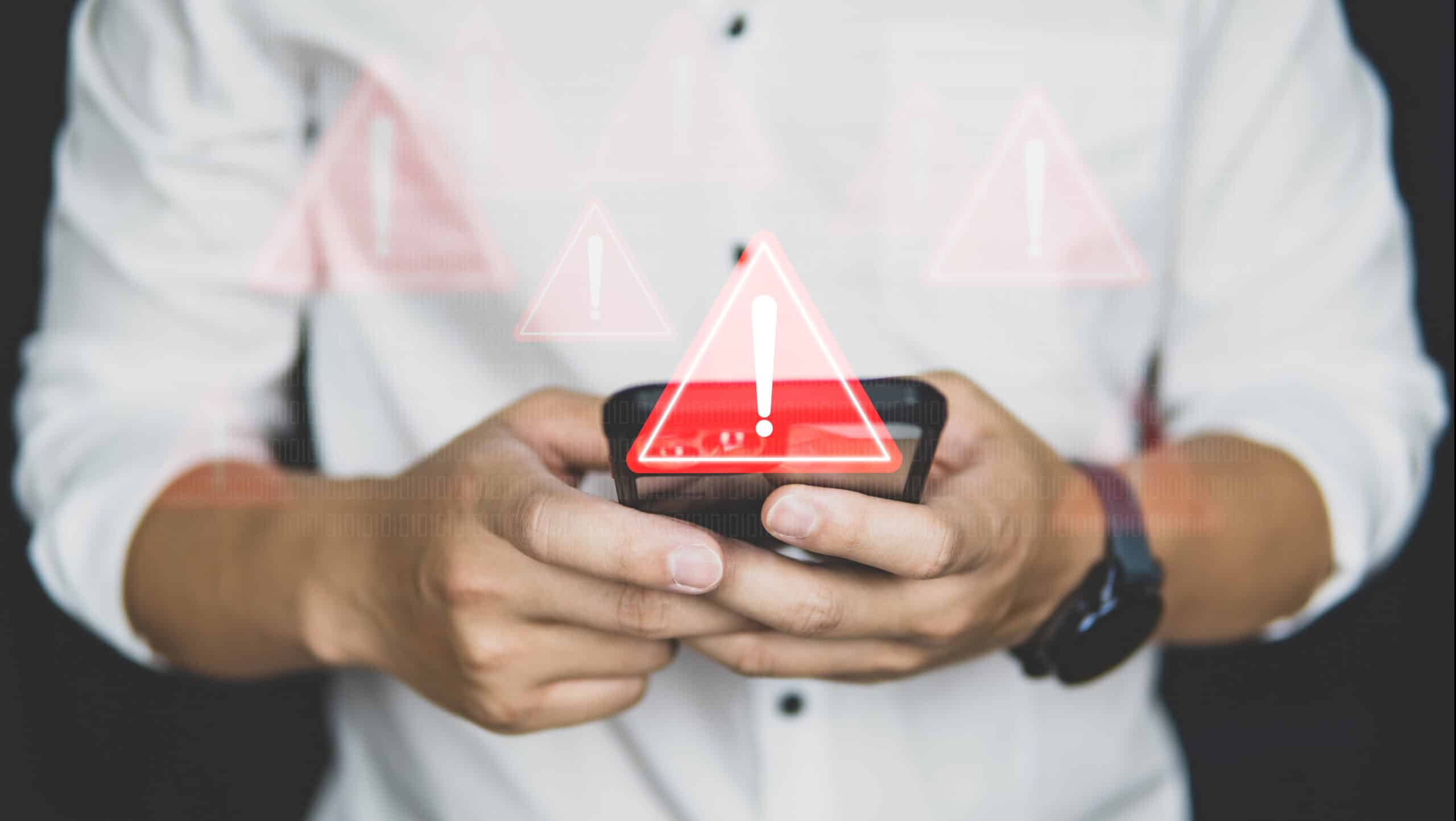A person in a white shirt holding a smartphone with red warning exclamation mark symbols displayed, suggesting alerts or notifications of potential danger or issues.