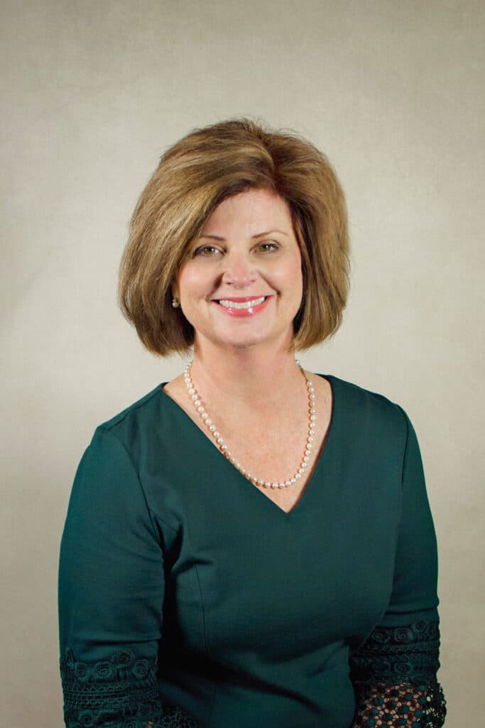 A woman with short light brown hair is smiling while seated in front of a plain beige background. She is wearing a dark green dress with lace sleeves and a pearl necklace.
